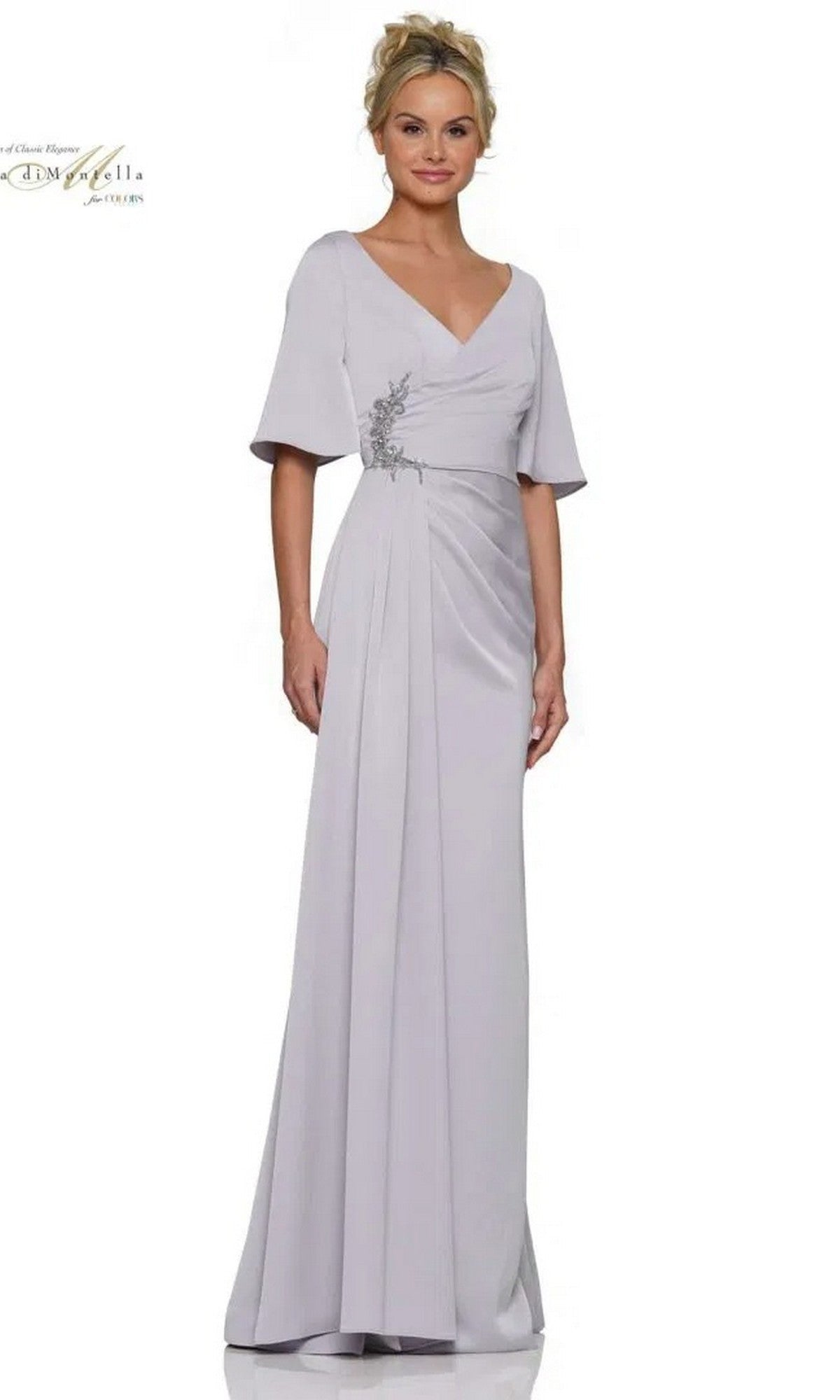 A woman models the Rina Di Montella RD2974 long formal dress in silver, featuring short sleeves, a V-neckline, ruched fitted bodice, pleated skirt, and floral waist detail, set against a white background.